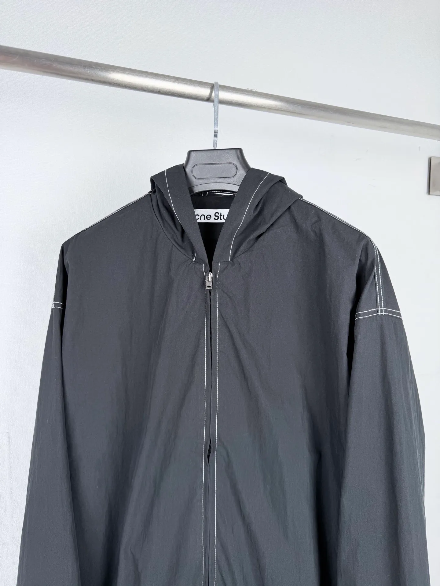 ACNE Studios back letter topstitch hooded jacket is made of fabric processing, contrasting topstitching, printing alignment and combination of elastic components. The fabric is treated with a physical wrinkle process, which does not rely on chemical coatings. It uses mechanical wrinkles to create a disordered texture effect on the surface of the fabric. While the fabric has a relaxed look, it still retains the structural tension of the woven fabric. During the cutting stage, the deformation caused by the folds of the fabric needs to be fully considered to ensure a stable silhouette after the pieces are spliced. The entire product uses contrasting topstitching as the main structural decoration. The seams of the cap, shoulder seams, cuffs, and hem are all exposed with contrasting topstitching. There are arc turns in different parts of the cut pieces. When sewing, it is necessary to control the fabric feeding rhythm and maintain a uniform stitch density. The elastic area of ​​the hem is sewn with multiple layers of materials. The layers of fabric, elastic bands, and hemming pieces are overlapped. It is easy for the line to shift and the fabric to arch, which requires high precision in the lathe operation. The Retro character printing on the back is the visual core of the entire garment. Multiple groups of characters are arranged in layers, giving it a worn and broken texture. The printing must ensure that the layer is strong and washable, but cannot cover up the details of the incomplete retro characters. At the same time, the printing positioning needs to be aligned with the middle seam of the back of the garment to avoid tilting and offset of the printing. The cuffs and hem are both elastic pleated structures. When sewing, you need to control the density of the fabric pleats to ensure a uniform and natural arc after shrinkage. The hood is spliced ​​into pieces and relies on multiple open threads to separate and shape it, ensuring that the hat is natural and three-dimensional and will not collapse against the face when worn. After all the sewing processes are completed, the garment needs to be ironed and finished to unify the wrinkles of the entire fabric and remove excess creases caused by the production process, finally presenting the complete Retro relaxed garment effect. Size: 46 48 50 52